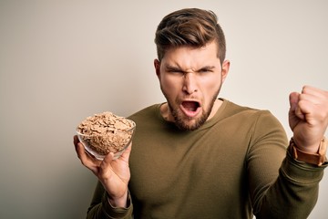 Young blond man with beard and blue eyes holding bowl with cornflakes cereals annoyed and frustrated shouting with anger, crazy and yelling with raised hand, anger concept