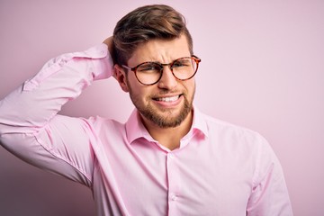 Young handsome blond man with beard and blue eyes wearing pink shirt and glasses confuse and wonder about question. Uncertain with doubt, thinking with hand on head. Pensive concept.