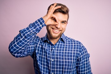 Obraz premium Young handsome blond man with beard and blue eyes wearing casual shirt standing doing ok gesture with hand smiling, eye looking through fingers with happy face.