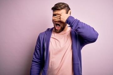 Young blond man with beard and blue eyes wearing purple sweatshirt over pink background peeking in shock covering face and eyes with hand, looking through fingers with embarrassed expression.