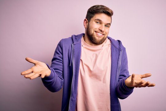Young blond man with beard and blue eyes wearing purple sweatshirt over pink background clueless and confused expression with arms and hands raised. Doubt concept.