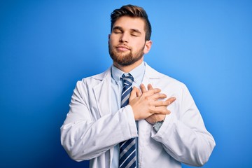 Young blond therapist man with beard and blue eyes wearing coat and tie over background smiling with hands on chest with closed eyes and grateful gesture on face. Health concept.