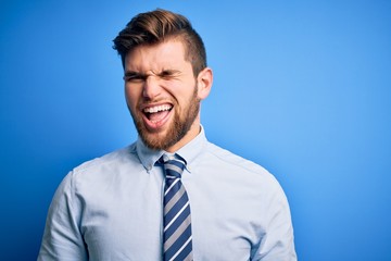 Young blond businessman with beard and blue eyes wearing elegant shirt and tie standing winking looking at the camera with sexy expression, cheerful and happy face.