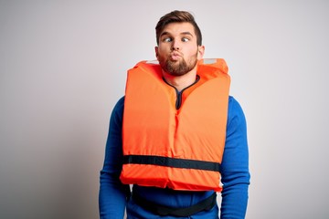 Fototapeta premium Young blond tourist man with beard and blue eyes wearing lifejacket over white background making fish face with lips, crazy and comical gesture. Funny expression.