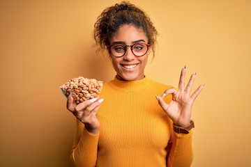 Young african american afro girl holding bowl with healthy peanuts over yellow background doing ok sign with fingers, excellent symbol