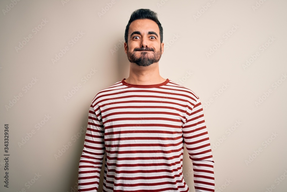 Poster Young handsome man with beard wearing casual striped t-shirt standing over white background puffing cheeks with funny face. Mouth inflated with air, crazy expression.