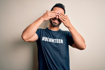Handsome man with beard wearing t-shirt with volunteer message over white background covering eyes...