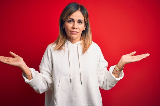 Middle age beautiful sportswoman wearing casual sweatshirt over isolated red background clueless and confused with open arms, no idea concept.
