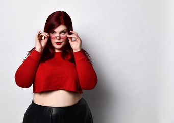 Overweight redhead woman in red spiked top, black skirt. She lowered her sunglasses and looking at you, posing isolated on white