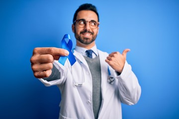 Young handsome doctor man with beard wearing stethoscope holding blue cancer ribbon pointing and showing with thumb up to the side with happy face smiling