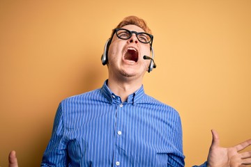 Young handsome redhead call center agent man wearing glasses working using headset crazy and mad...