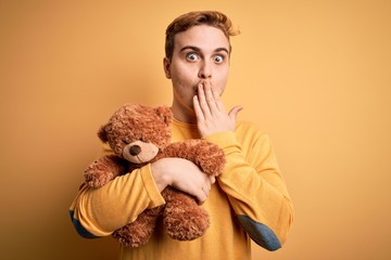 Young handsome redhead man holding teddy bear animal toy over yellow background covering mouth with...