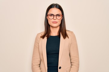 Young beautiful brunette businesswoman wearing jacket and glasses over white background skeptic and...