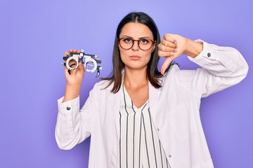 Young beautiful brunette oculist woman holding optometry glasses over purple background with angry face, negative sign showing dislike with thumbs down, rejection concept