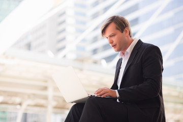 An adult, Ukrainian businessman wearing an official dress, typing a document in a laptop, having a building background and outdoor structure.