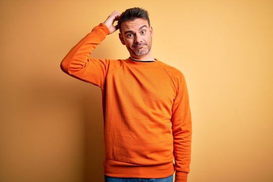 Young handsome man wearing orange casual sweater standing over isolated yellow background confuse and wonder about question. Uncertain with doubt, thinking with hand on head. Pensive concept.