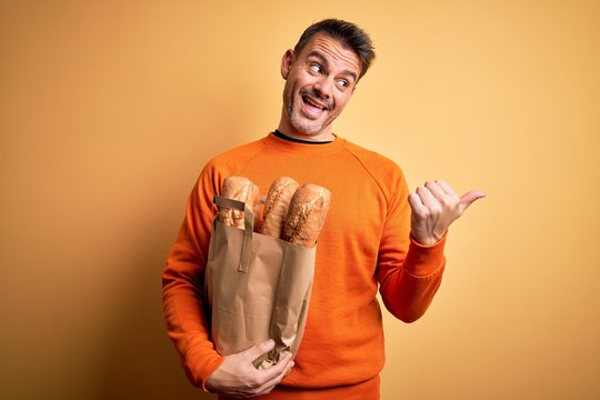 Young handsome man holding paper bag with bread over isolated yellow background pointing and showing with thumb up to the side with happy face smiling