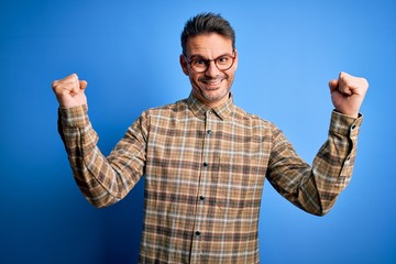 Naklejka premium Young handsome man wearing casual shirt and glasses standing over blue background celebrating surprised and amazed for success with arms raised and open eyes. Winner concept.