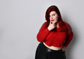 Obese redhead lady in red spiked top, black leather skirt, earrings. Touching face, put hand on waist, posing isolated on white