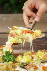 Hand holding a slice of Pizza with Mozzarella cheese, Ham, Tomatoes, salami, pepper, pepperoni. Pizza on wooden background.