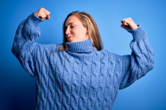 Young beautiful blonde woman wearing casual turtleneck sweater over blue background showing arms muscles smiling proud. Fitness concept.