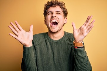 Young blond handsome man with curly hair wearing green sweater over yellow background celebrating mad and crazy for success with arms raised and closed eyes screaming excited. Winner concept