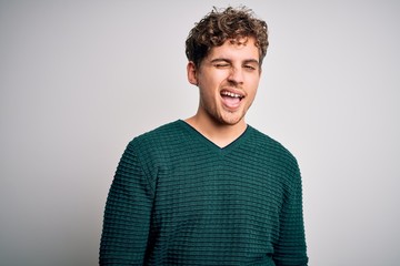 Young blond handsome man with curly hair wearing green sweater over white background winking...