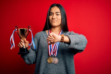 Young beautiful champion asian woman holding trophy wearing medals over red background with angry...