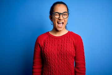 Young beautiful asian woman wearing casual sweater and glasses over blue background winking looking...