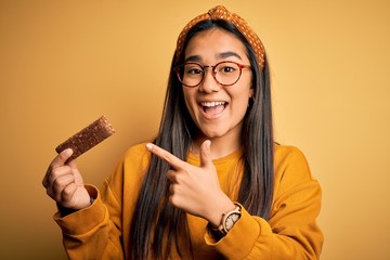 Young beautiful asian woman eating healthy protein bar over isolated yellow background very happy pointing with hand and finger
