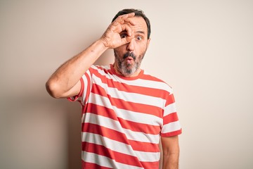 Middle age hoary man wearing casual striped t-shirt standing over isolated white background doing ok gesture shocked with surprised face, eye looking through fingers. Unbelieving expression.