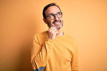 Middle age hoary man wearing casual sweater and glasses over isolated yellow background with hand on chin thinking about question, pensive expression. Smiling and thoughtful face. Doubt concept.