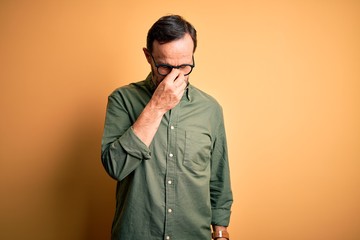 Fototapeta premium Middle age hoary man wearing casual green shirt and glasses over isolated yellow background tired rubbing nose and eyes feeling fatigue and headache. Stress and frustration concept.