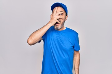 Middle age handsome deliveryman wearing cap standing over isolated white background peeking in shock covering face and eyes with hand, looking through fingers afraid