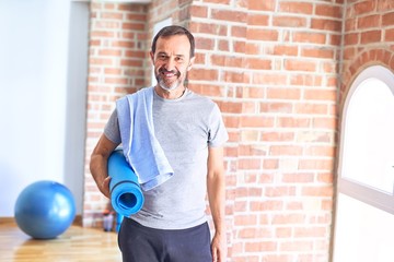 Middle age handsome sportman holding mat and towel standing before do exercise at gym with a happy...