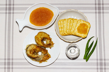 Fried fish in a white plate, caviar in a porcelain plate in the form of a fish, a glass of vodka, crackers with caviar and green onions on a white tablecloth. Appetizing fish appetizer for one.