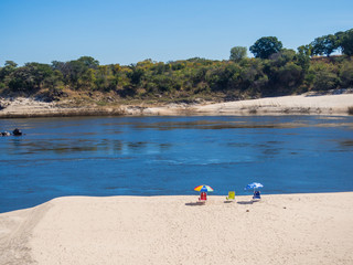 Three Sun Loungers With Umbrellas On Deserted Beach Of River Zambezi Against Clear Sky, Zambia