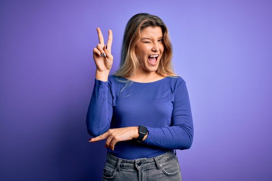 Young beautiful blonde woman wearing casual t-shirt over isolated purple background smiling with happy face winking at the camera doing victory sign with fingers. Number two.