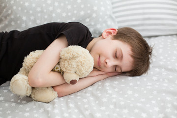 a boy sleeping holding toy teddy bear. Boy pretending to be sleeping in daylight. Bedroom with light gray bed linen. Little boy holds his teddy bear. Stay at home concept. Family time at home. 