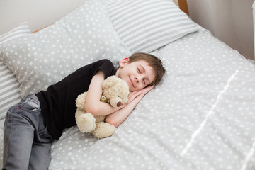 a boy sleeping holding toy teddy bear. Boy pretending to be sleeping in daylight. Bedroom with light gray bed linen. Little boy holds his teddy bear. Stay at home concept. Family time at home. 