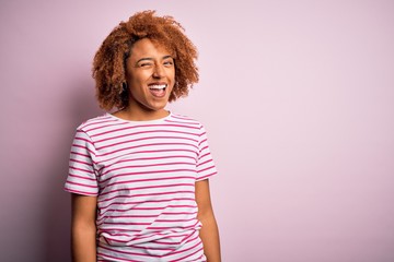 Young beautiful African American afro woman with curly hair wearing casual striped t-shirt winking...