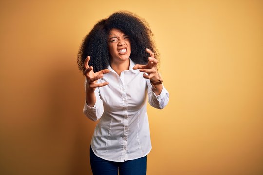 Young beautiful african american elegant woman with afro hair standing over yellow background Shouting frustrated with rage, hands trying to strangle, yelling mad