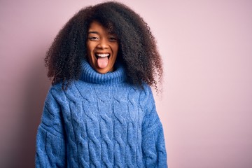 Young beautiful african american woman with afro hair wearing winter sweater over pink background sticking tongue out happy with funny expression. Emotion concept.