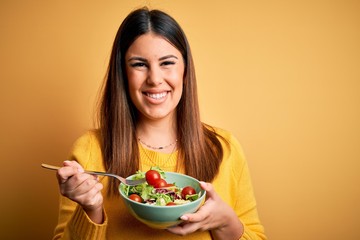 Young beautiful woman eating healthy fresh salad over yellow background with a happy face standing...