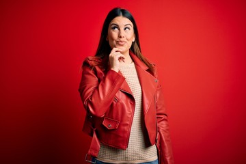 Young beautiful woman wearing red leather jacket over isolated background with hand on chin thinking about question, pensive expression. Smiling with thoughtful face. Doubt concept.