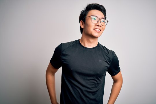 Young handsome chinese man wearing black t-shirt and glasses over white background looking away to side with smile on face, natural expression. Laughing confident.