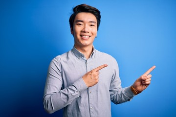 Young handsome chinese man wearing casual shirt standing over isolated blue background smiling and looking at the camera pointing with two hands and fingers to the side.