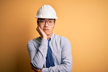 Young handsome chinese architect man wearing safety helmet and tie over yellow background thinking looking tired and bored with depression problems with crossed arms.