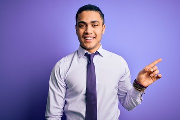 Young brazilian businessman wearing elegant tie standing over isolated purple background with a big smile on face, pointing with hand finger to the side looking at the camera.
