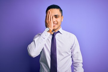 Young brazilian businessman wearing elegant tie standing over isolated purple background covering one eye with hand, confident smile on face and surprise emotion.